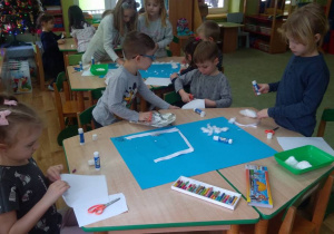 Dzieci przy stoliku wycinają kawałki białej kartki i naklejają na niebieskim kartonie.